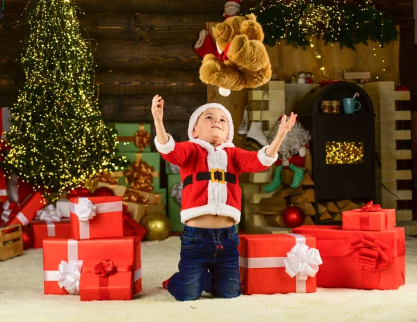 見てみろよ。息子は冬休みを祝う準備ができてる。小さな男の子が興奮してプレゼント。子供は熊のおもちゃの贈り物を投げる。Xmasにおもちゃ屋がオープンしました。メリー・クリスマス。新年明けましておめでとう。ハッピーチャイルドサンタ帽子 — ストック写真
