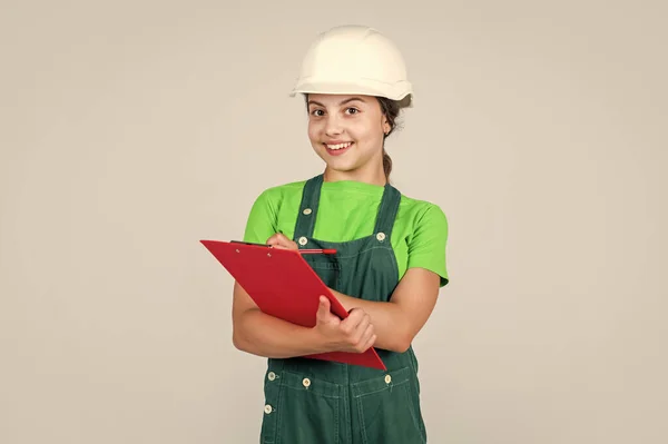 En liten unge i hjälm med surfplatta. Reparationsplanering. Grabben i hatt gör anteckningar. Dokumentmapp för barn. Ungen lär sig att vara en byggarbetare byggare med säkerhetshatt — Stockfoto