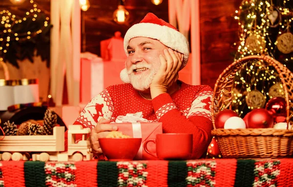 Ano Novo. Uma casa acolhedora. Bebida quente e biscoitos. Véspera de Natal. Atmosfera alegre. Um homem alegre, o Pai Natal, a beber chá. Férias de inverno. Avô alegre com presente. Avô sonhador em chapéu de Santa — Fotografia de Stock