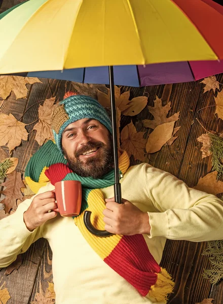 Fröhlicher Mann, der heißen Tee oder Kaffee trinkt. Herbstliche Rabatte auf Kleidung. Mann mit Regenbogen-Regenschirm. Er mag Regen. Der Wetterbericht verspricht Regen. Bärtiger Mann mit buntem Regenschirm — Stockfoto