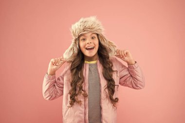Sıcak bir gülümseme. Mutlu çocuk pembe arka planı. Soğuk mevsim için sıcak kıyafetler. Çocuk modası. Moda kızı hipster 'a benziyor. Sonbahar tarzı. Çocukluk aktivitesi. Tüylü kulaklıklı şapka aksesuarı. Küçük kız kış şapkası