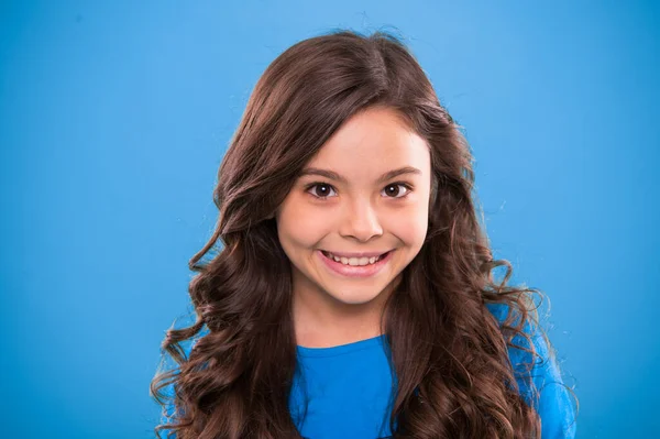 Cabelo de ondulação perfeito. Miúdo menina longo cabelo brilhante saudável. Kid rosto bonito feliz com penteado encaracolado adorável ficar sobre fundo azul. Ensinar hábitos saudáveis de cuidado do cabelo. Menina crescer cabelos longos — Fotografia de Stock