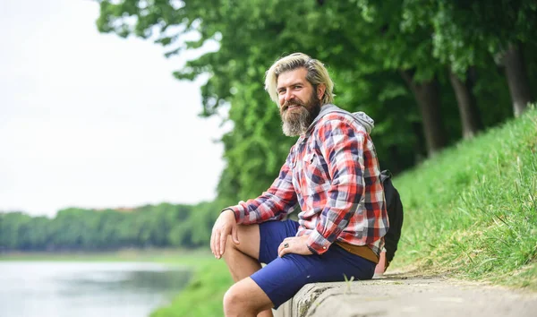 Nice day. Rest and relax. Relaxation and pleasure. Bearded hipster relax outdoors. Man tourist sit riverside. Man with beard and moustache relaxing nature background. Tourism concept. Summer vacation