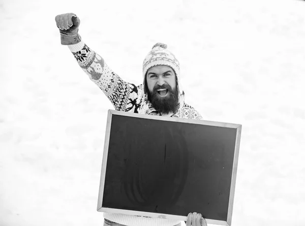 Winter event. Man with beard hold chalkboard copy space. Check it out. Winter holidays. Emotional guy snowy nature background. Winter announcement. Hipster knitted hat and gloves show blackboard