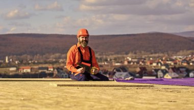 Nitelikli inşaatçı çatı döşemesi kuruyor. kopyalama alanı. Çatı keçesi için inşaat işi. Çatı endüstrisi. Çatı müteahhidi koruyucu miğferiyle çatıda yatıyor. Çatı tamircisi hasar görmüş çatıyı inceliyor.