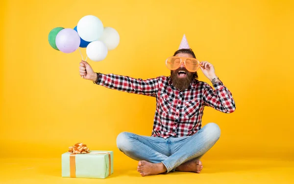 Ο χαλαρός τύπος με τα χαρούμενα γενέθλια φαίνεται χαρούμενος. γενειοφόρος άνθρωπος αισθάνεται τη χαρά. άνθρωπος με κουτί δώρου γιορτάζει τα γενέθλια. ώριμος hipster με φωτεινά πολύχρωμα μπαλόνια. Τρελός αστείος τύπος — Φωτογραφία Αρχείου