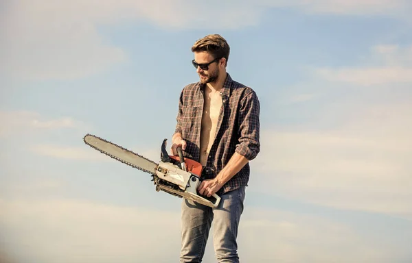 Handsome man with chainsaw blue sky background. Gardener lumberjack equipment. Lumberjack with chainsaw in his hands. Dangerous job. Feeling confident. Risky temper. Powerful chainsaw. Sharp blade