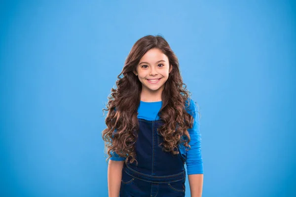 Extreme hair volume. Kid girl long healthy shiny hair. Kid happy cute face with adorable curly hairstyle stand over blue background. Little girl grow long hair. Teaching healthy hair care habits
