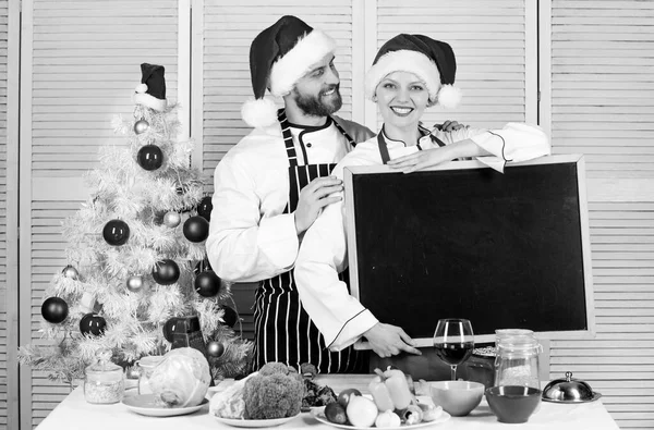 Menu voor onze familie. Kerstmaaltijd aan het koken. Man en vrouw chef-kok santa hoed in de buurt van kerstboom houden schoolbord kopieerruimte. Kerstrecept concept. Traditioneel familiemenu voor kerstdiner — Stockfoto
