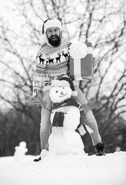 Surprise concept. Winter games. Winter activity. Winter vacation. Guy happy face snowy nature background. Hipster with beard hold gift box. Man made snowman. Man Santa hat having fun outdoors