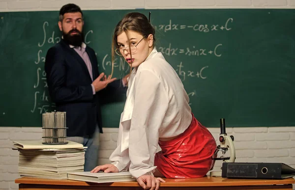 同僚といちゃつく。誰もがそんな先生を夢見ている。レザースカートの魅力的な教師。生意気な先生。突然の学生。科学はセクシーだ。セクシーな女の子座っていますテーブル一方、男スタンド黒板 — ストック写真