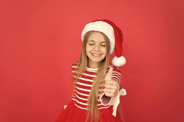 新年に向けて親指を立てる。新年のサンタの表情で幸せな女の子の笑顔。小さな子供がジェスチャー赤の背景を親指を表示します。新年会。新年明けましておめでとう。メリークリスマス — ストック写真