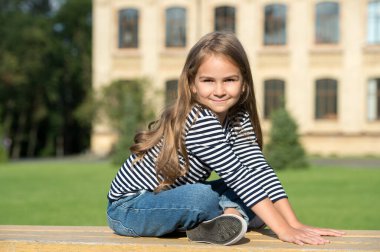 Gündelik moda tarzında uzun saçlı mutlu küçük çocuk güneşli bir yaz gününde bankta oturup dinleniyor.