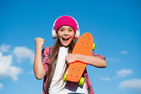 ハッピー女の子作る勝者ジェスチャー保持スケートボードと現代のヘッドフォンで音楽を聴く晴れ空屋外、スケートボード — ストック写真