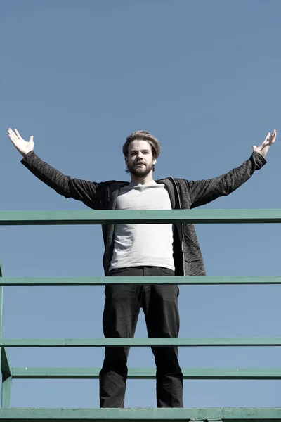 Chico de pie en la terraza de la azotea en el cielo azul. Hombre barbudo con las manos levantadas en el exterior soleado. Éxito y concepto futuro. Moda de hombre, estilo casual — Foto de Stock