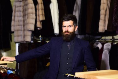 Katı yüzlü adam arka planda kürklü paltoların yanında oturuyor..