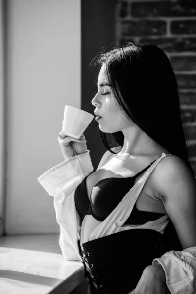 Насолоджуйтесь ранковим сонячним світлом. Чуттєва дівчина сексуальні груди п'є каву біля підвіконня. Приваблива жіноча білизна розслабляється вдома. Пристрасний таємничий коханець. Ідеальний час для кави. Але перша кава — стокове фото
