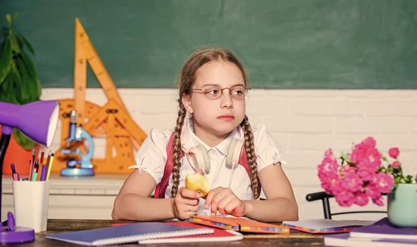 Okul hayatı konsepti. Modern çocukluk. Okul tatili. Liseli kız masa başında tahtada oturuyor. Okuldaki çocuk öğrenci. Elma meyvesi yiyen küçük bir kız. Sağlıklı bir yaşam tarzı. Ders aralarında atıştırmalık