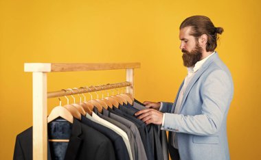 Moda bloguna devam. Erkek giyimi konsepti. Adam giyim mağazasına alışverişe gider. Stüdyo atölyemiz herhangi bir moda tarzını sunar. Bir sürü erkek giysisi. Sakallı adam askıda ceketi seçer.