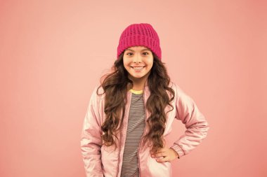 Sıcak kış kıyafetleri içinde neşeli bir çocuk. Çocuklar için mevsimlik moda. Küçük pembe duvar. Küçük kız kabarık ceket ve örgü şapka. Soğuk havada sağlık hizmeti. Havası nasıl olursa olsun