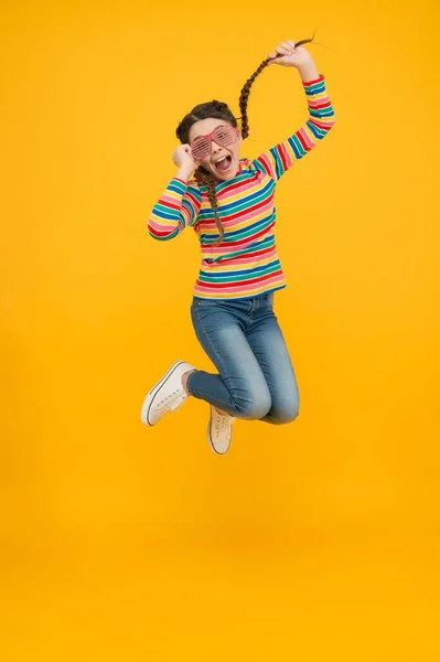 Menina criança salto feliz com cabelo trançado usar cor do arco-íris e óculos de festa glamour no fundo amarelo, extático — Fotografia de Stock