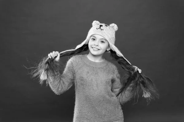 Boldogság és öröm. Téli ruha. Kis gyermek hosszú haj viselni kalap piros háttér. Aranyos modell élvezni téli stílusban. Imádnivaló kis medve. A kisgyerek kötött sapkát visel. Kislány téli divat tartozék — Stock Fotó