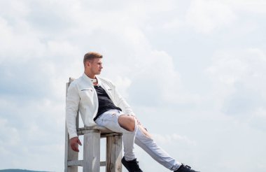 Gündelik tarzda giyinen yakışıklı genç adam tahta koltukta rahatla temiz havanın tadını çıkar gökyüzü bulutlu, fotokopi alanı için dinlen.