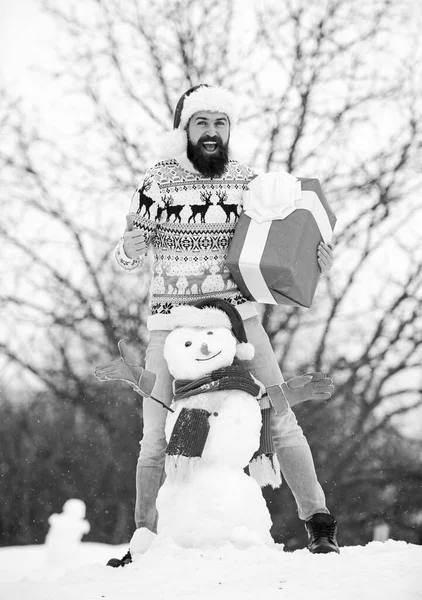 Man made snowman. Man Santa hat having fun outdoors. Guy happy face snowy nature background. Hipster with beard hold gift box. Surprise concept. Winter games. Winter activity. Winter vacation