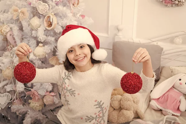 Fancy decor. Creating festive atmosphere. Child decorating christmas tree with red ball. Girl kid decorating christmas tree. Cherished holiday activity. Kid in santa hat decorating christmas tree — Stock Photo, Image