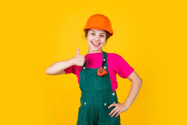 Kid wear helmet on construction site. teen girl builder with building tool tape-measure. child on repairing work. concept of renovation in workshop. busy professional carpenter show thumb up — Stock Photo, Image