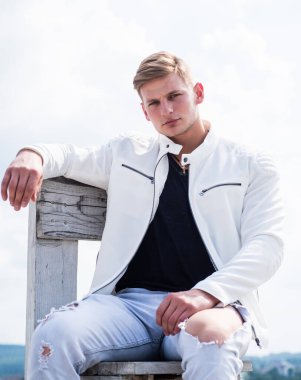 Yakışıklı, gündelik tarzda genç bir adam ahşap sandalyeye oturur açık havada bulutlu gökyüzü dalgın bir görünüm, ciddi.