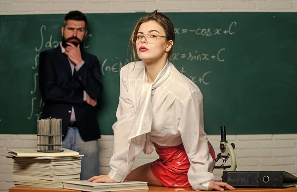 Attractive teacher in leather skirt. Cheeky teacher. Impudent student. Science is sexy. Sexy girl sit table while man stand chalkboard. Flirting with colleague. Everyone dreaming about such teacher