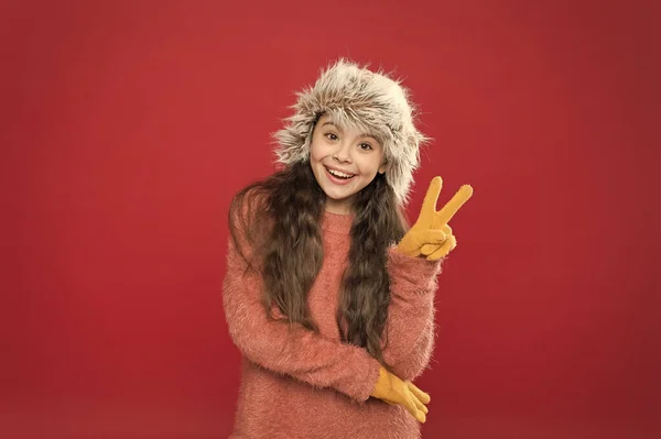 Béke mindenkivel. Gyermek hosszú haj puha kalap. Téli divat koncepció. Meleg kalap a hideg téli időjáráshoz. Gyerek lány mosoly piros háttér. Puha szőrös kiegészítő. Téli szezon. Kis divatbemutató koncepció — Stock Fotó