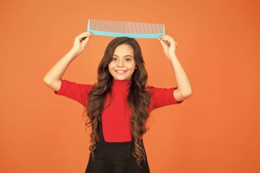 Kocaman tarağı olan neşeli, gülümseyen küçük çocuk. Kuaför salonu. Savaş saçı. Moda ipuçları. Karışık saç için saç fırçası. Eğleniyorum. Mükemmel saçlar. Geniş tarak. Muhteşem uzun saçlı kız, büyük tarak tutuyor.