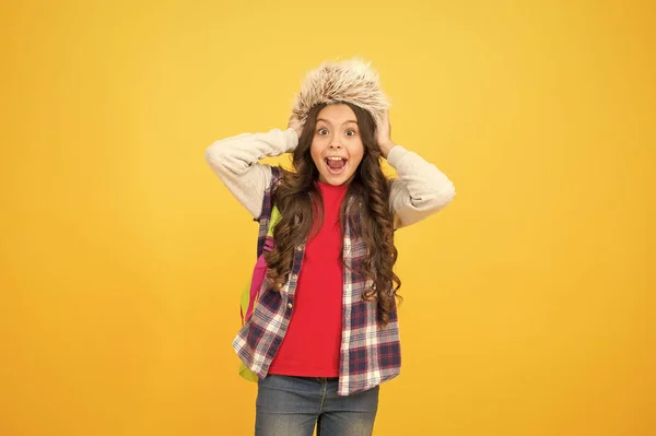 Che grande sorpresa. pupilla spensierata. piccola ragazza indossare cappello auricolare. periodo scolastico invernale e vacanze. corsi invernali di educazione. zaino studentessa sfondo giallo. periodo di selezione del corso — Foto Stock