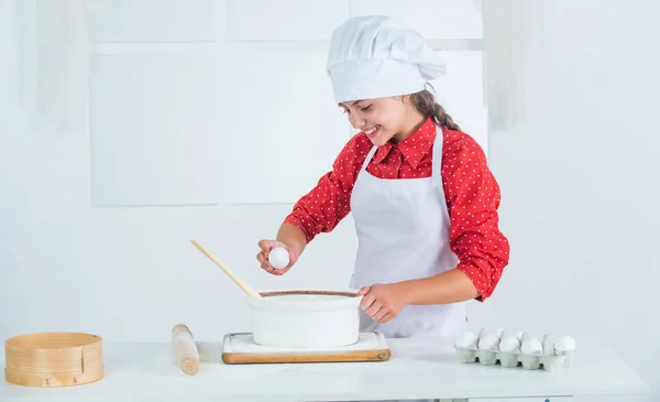 Das Ei hinzufügen. Zeit zum Essen. glückliches Kind, das in der Küche kocht. Plätzchen backen in der Küche. professionelle und qualifizierte Bäcker. Kind in Kochuniform und Hut. Teenie-Mädchen bereitet Teig zu. Kuchen nach Rezept zubereiten — Stockfoto