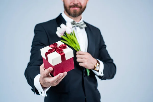 Barbudo hombre vista recortada celebrar caja de regalo atada con lazo de cinta roja enfoque selectivo y flores de primavera para la celebración fondo gris, 8 de marzo — Foto de Stock