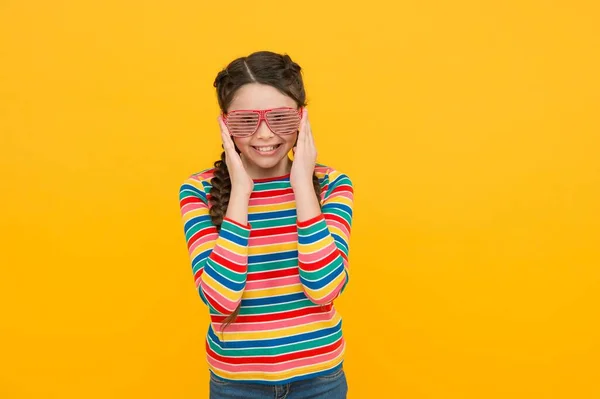 Glücklich Kind Mädchen mit geflochtenen Haaren tragen Regenbogenfarbe und Glamour-Party-Brille auf gelbem Hintergrund, Spaß Zeit — Stockfoto