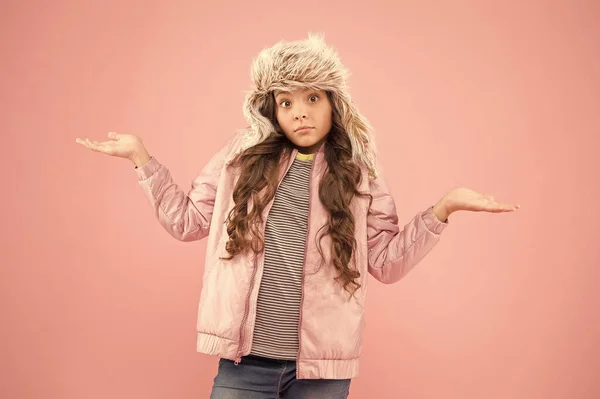 at a loss. warm clothes for cold season. fur hat accessory. small girl winter earflap hat. bewildered child pink background. kid fashion. girl look like hipster. autumn style. Childhood activity