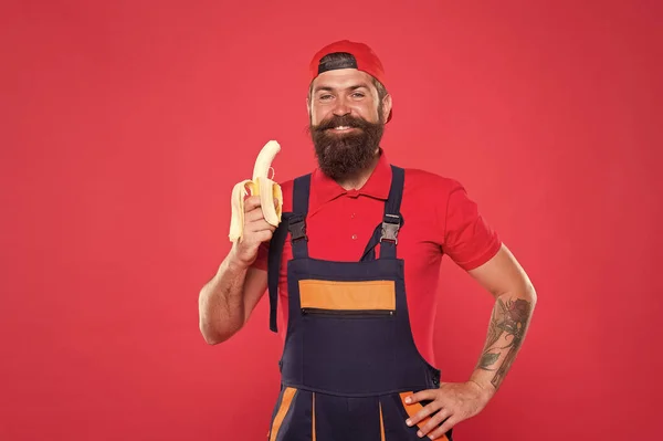 Faça um lanche saudável. Homem barbudo de uniforme e gorro comendo banana. Trabalhador construtor mecânico comer banana. Fruta fresca. Viver uma vida saudável. Conceito saboroso e saboroso. Morde banana. Handyman com fome — Fotografia de Stock