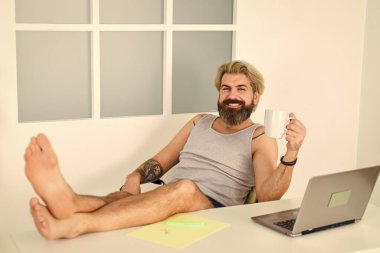 Sakallı adam kahve içer ve rahatça çalışır. İnternette kişisel eğitim. Evde kal. Koronavirüs karantinasına kilitlenin. Uzak bir iş. Yalın ayaklı işadamı merkez ofiste dizüstü bilgisayarda çalışıyor.