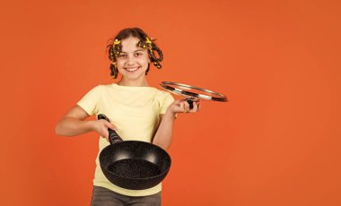 Tavalı şirin küçük kız portresi. Tavalı komik bir ev hanımı. Bukle makinesi ve tavası olan sevimli, komik bir kız. Küçük kız ev hanımı mutfakta yemek pişiriyor.