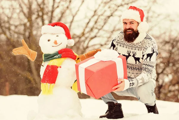 冬休み。男は雪だるまを作った。サンタさんの帽子は屋外で楽しんでいます。男幸せな顔雪の性質の背景。髭を生やしたヒップスターはギフトボックスを保持します。驚きのコンセプト。冬の試合だ。冬のアクティビティ — ストック写真
