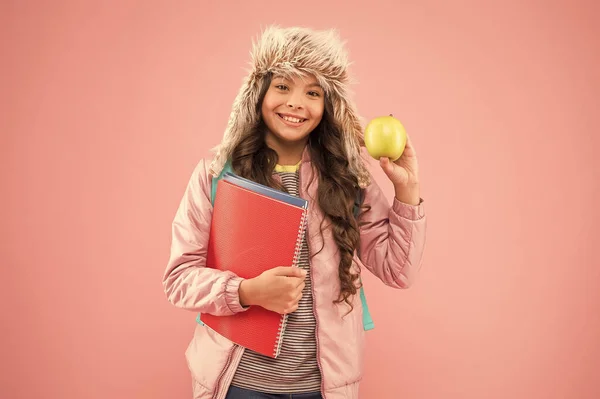 Schulmädchen mit Büchern nach dem Unterricht. Schulpause. kleine glückliche Mädchen Ohrenklappen Hut Apfel essen. Winterurlaub. Teenager-Kind rosa Hintergrund. Schüler-Alltag. Moderne Bildung. Student des Monats — Stockfoto