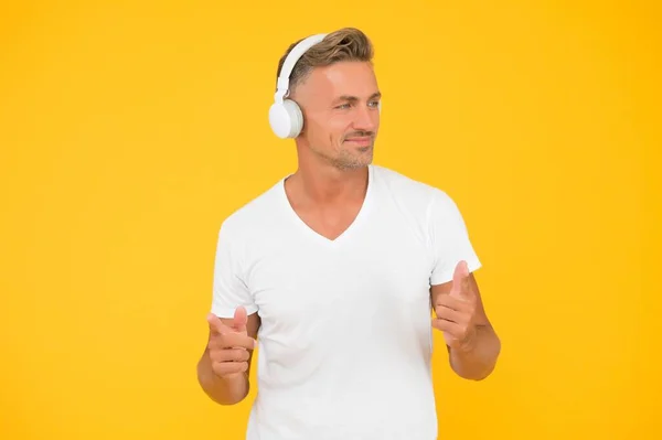 Happy middle-aged man listen to music in modern headphones pointing index fingers yellow background, promoting — Stock Photo, Image