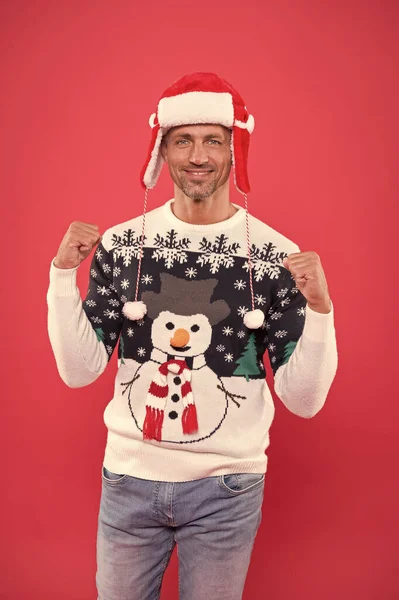 unshaven man funny sweater red background. male cold weather fashion. santa man ready for winter holiday. xmas party and activity. christmas is here. happy new year. sexy man santa earflap hat