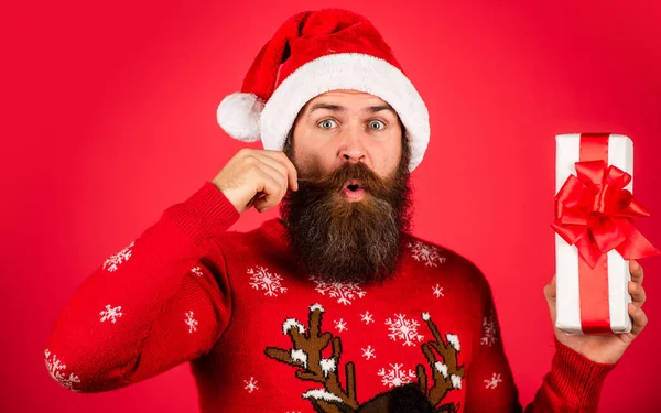 Rolničky. zimní prázdninová párty. dospělý muž slaví nový rok. vousatý hipster ve vánočním svetru. překvapený muž s vousy v klobouku Santa Clause. brutální muž drží dárkovou krabici. vánoční online nakupování — Stock fotografie