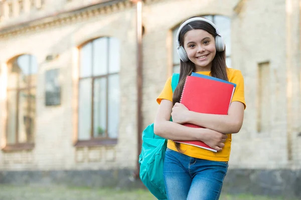 Glückliches Kind trägt Schultasche und Buch mit modernen Kopfhörern zum Zuhören, Kopierraum — Stockfoto
