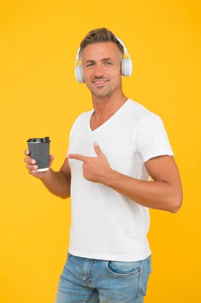 Glücklicher Mann hört Musik in modernen Kopfhörern und zeigt mit dem Finger auf Kaffeetasse zum Mitnehmen gelben Hintergrund, um zu bestellen — Stockfoto