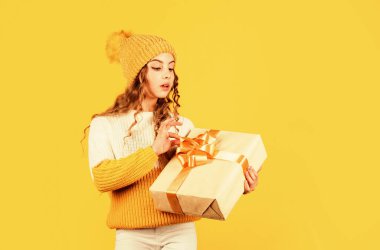 Noel partisi zamanı. Neşeli küçük kız örgü şapka ve kazak örüyor. Çocuk hediye kutusunu sarı arka planda tutuyor. Xmas hediye alışverişi. Noel 'in aydınlık olsun. Mutlu kış tatilleri.